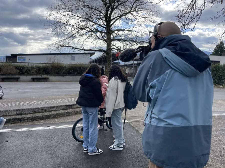 Marc filme deux collégiennes observant une troisième camarade faisant du vélo. Les deux collégiennes sont de dos. Elles portent leurs sacs à dos à l'épaule et manteau pour se protéger du froid de l'hiver.