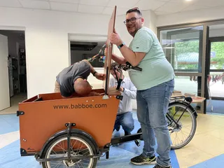 Trois hommes réparent la caisse avant d'un vélo cargo. Un des hommes regarde la caméra tandis que les deux autres sont affairés